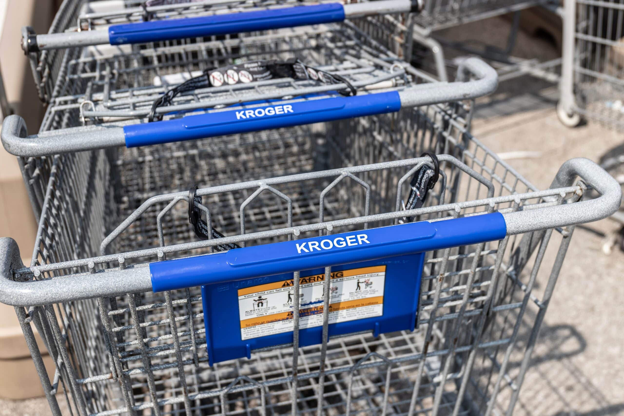 Rows of Kroger shopping carts lined up outside a grocery store, featuring blue handles with the Kroger brand name visible.