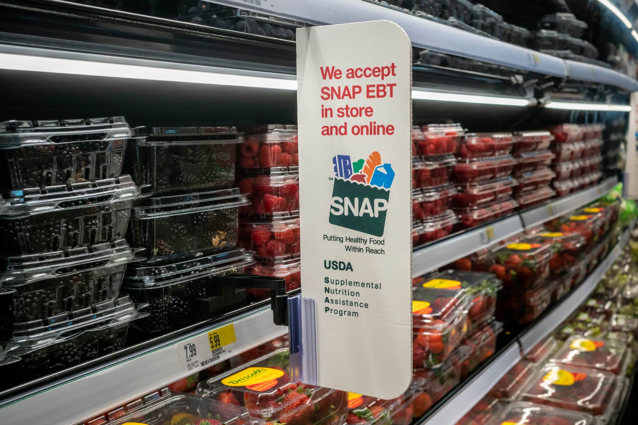 A SNAP EBT assistance sign stands in a grocery store produce section, highlighting food assistance access for shoppers.