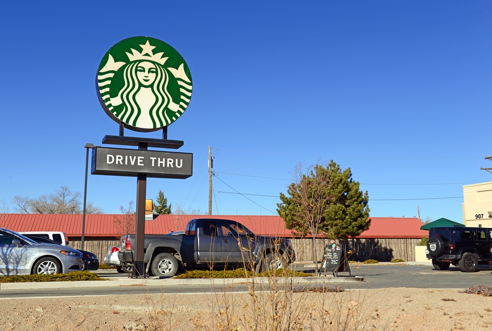 Starbucks drive-thru sign outside a coffee shop with parked vehicles under a clear blue sky.