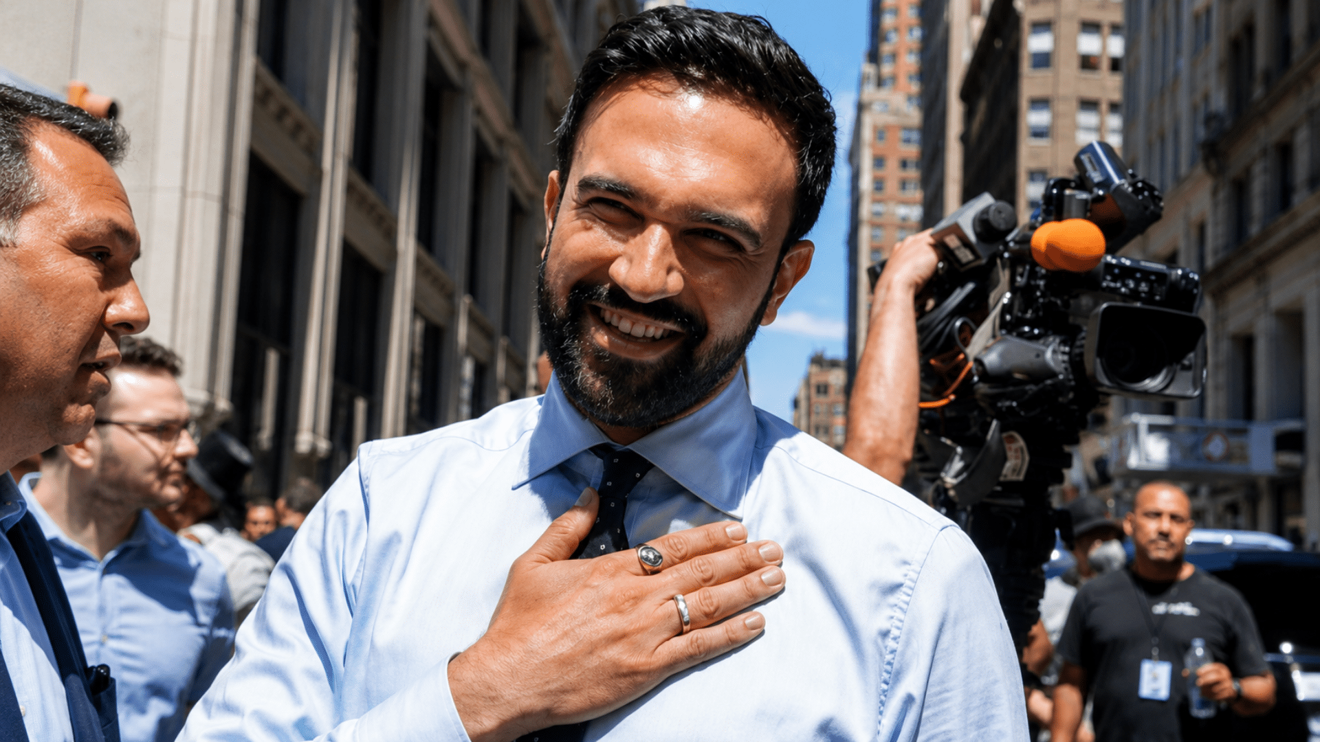 Zohran Mamdani smiling in a light blue dress shirt and tie with his hand over his heart amidst a crowd and media cameras.