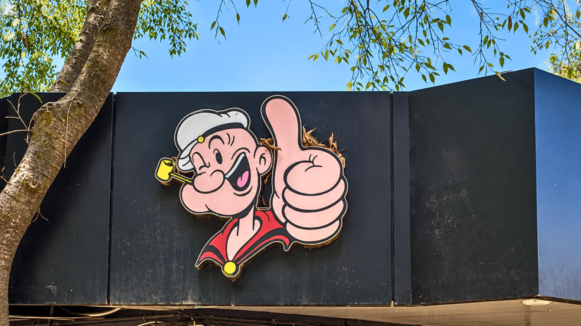 A Popeyes restaurant exterior sign featuring the classic cartoon character Popeye the Sailor giving a thumbs up.