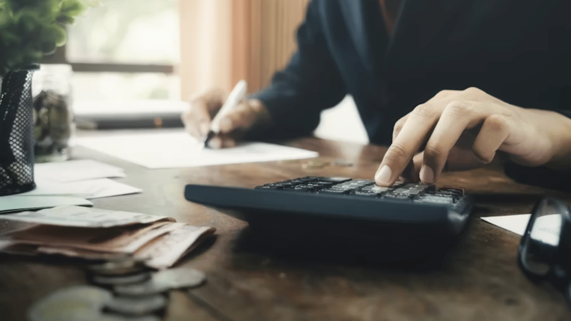 A person in a business suit using a calculator at a wooden office desk with paper money and coins nearby.