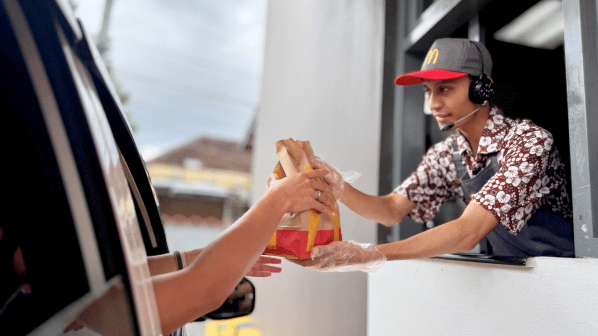 A McDonald's employee wearing a headset and gloves handing a paper takeout bag through a drive-thru window to a customer.