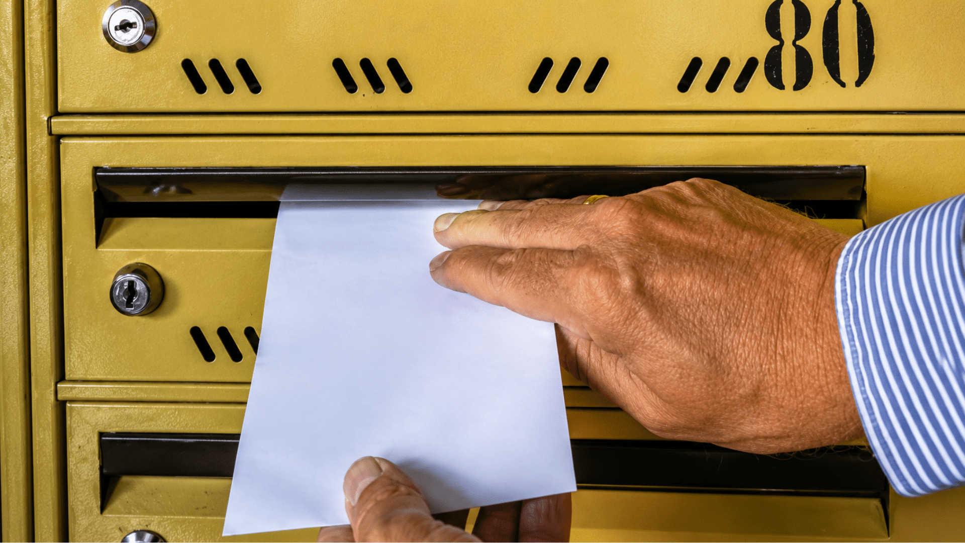 A close-up shot of a person's hands sliding a white envelope into a yellow metal mailbox slot.