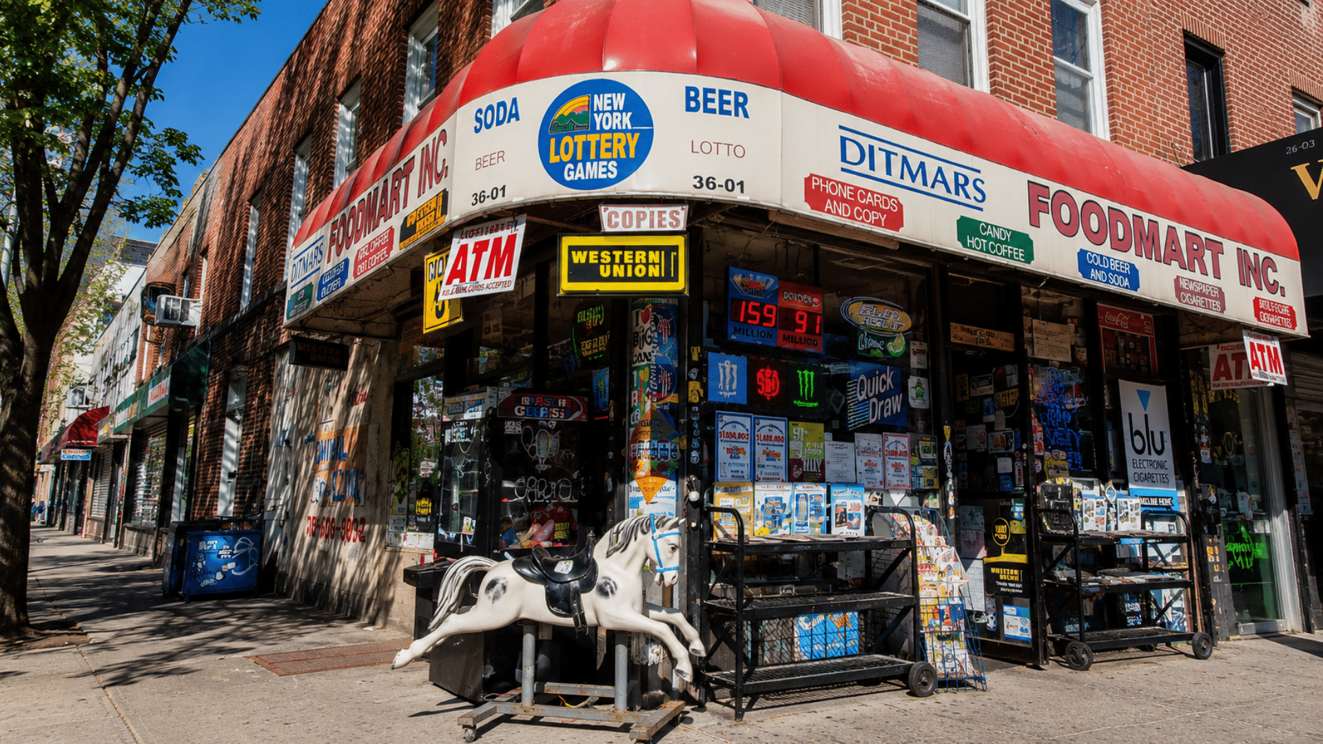 A street corner bodega storefront under a red awning displaying signs for lottery games and ATM services.