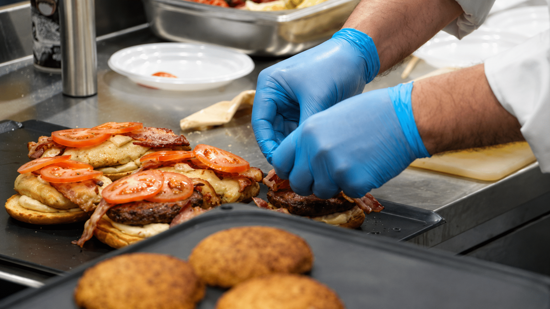 A kitchen worker wearing blue gloves assembling fresh burgers with bacon and sliced tomatoes on a counter.