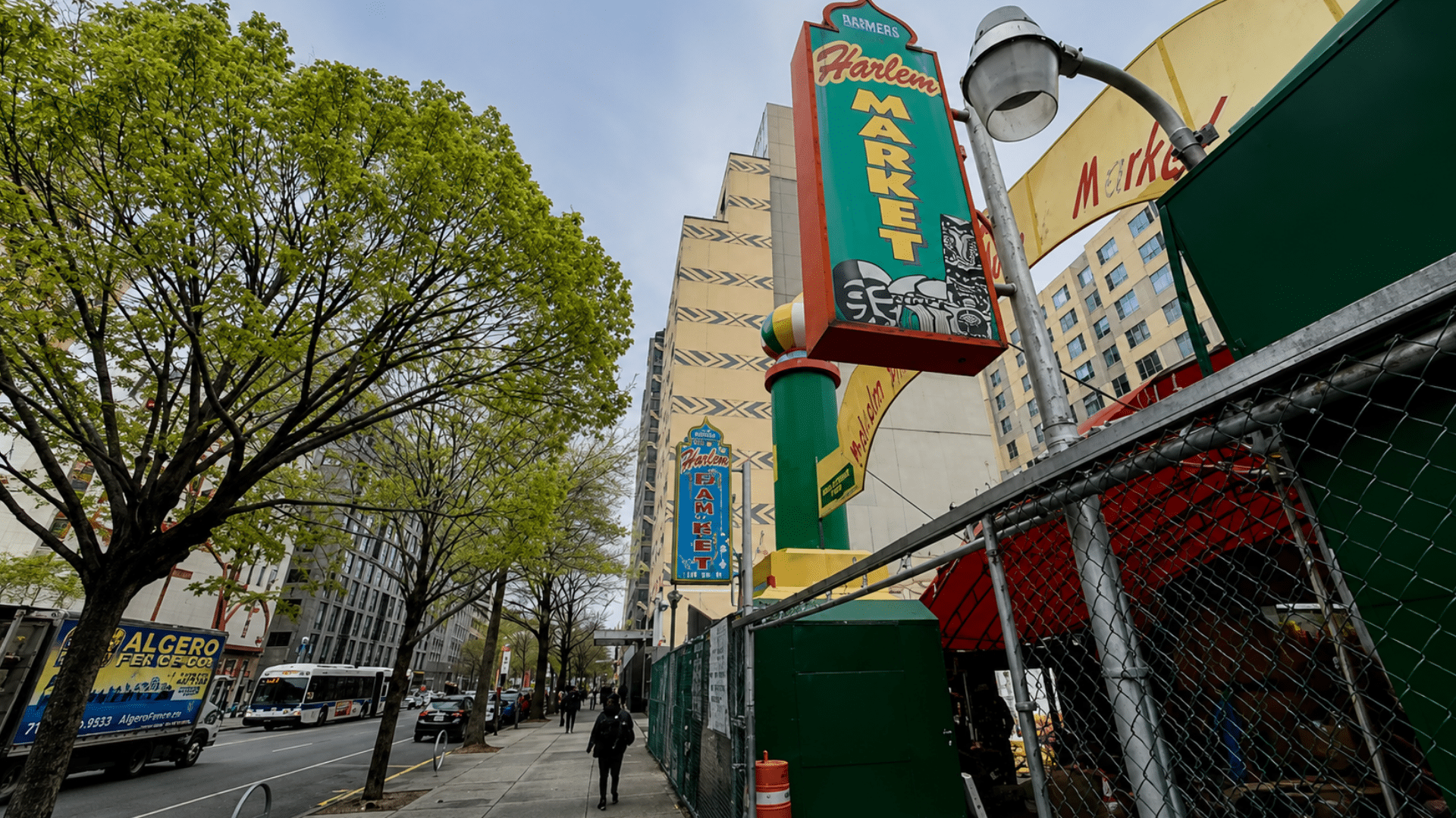 A person walking along a city sidewalk in front of the Harlem Market storefront sign.