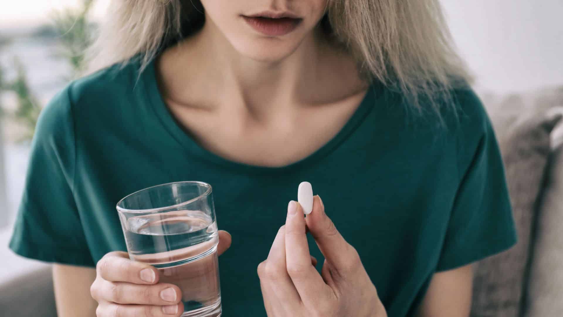 Woman holding a glass of water and a white tablet, preparing to take medication.