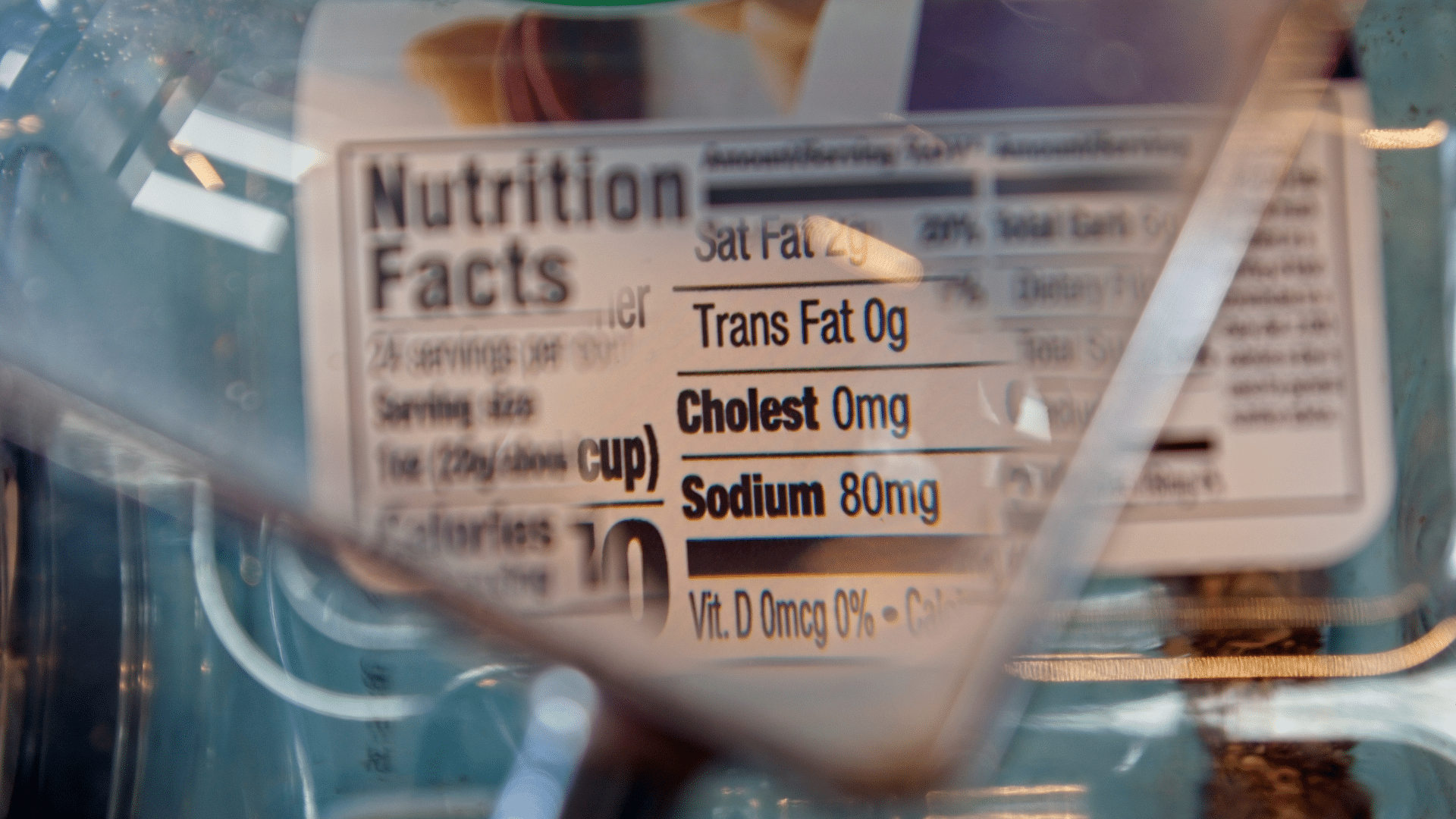 A close-up view through a plastic surface magnifies a _Nutrition Facts_ panel, highlighting saturated fat, trans fat, cholesterol, and sodium metrics.