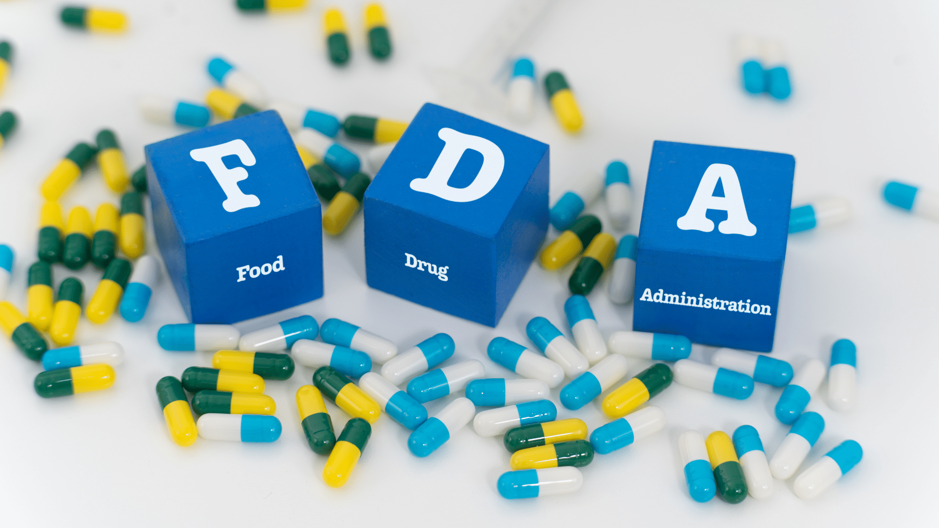 Blue toy blocks spelling out _FDA_ and _Food Drug Administration_ sit arranged over a scattered pile of green, yellow, and white medicine capsules.