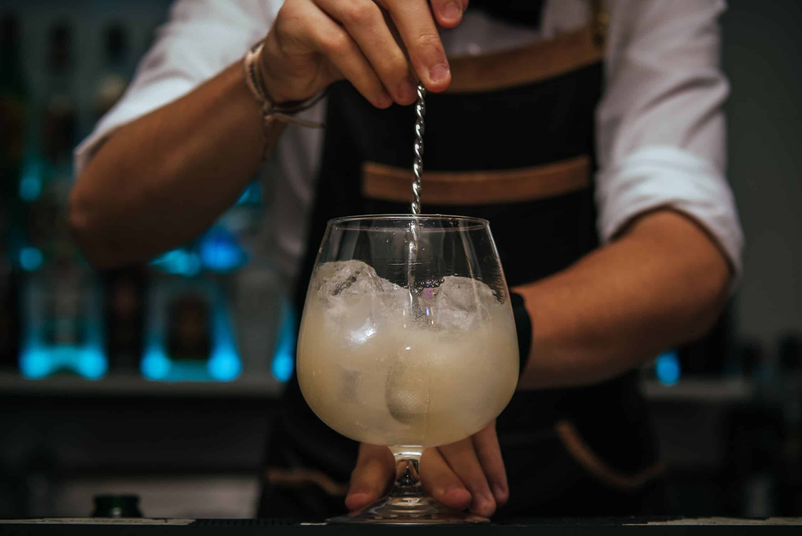 Bartender stirring a cocktail with ice in a stemmed glass behind a bar.