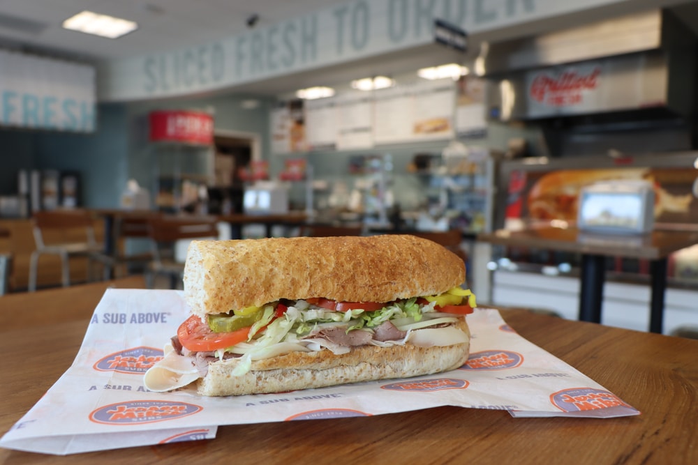A Jersey Mike’s sub sandwich sits on a table inside a restaurant dining area.