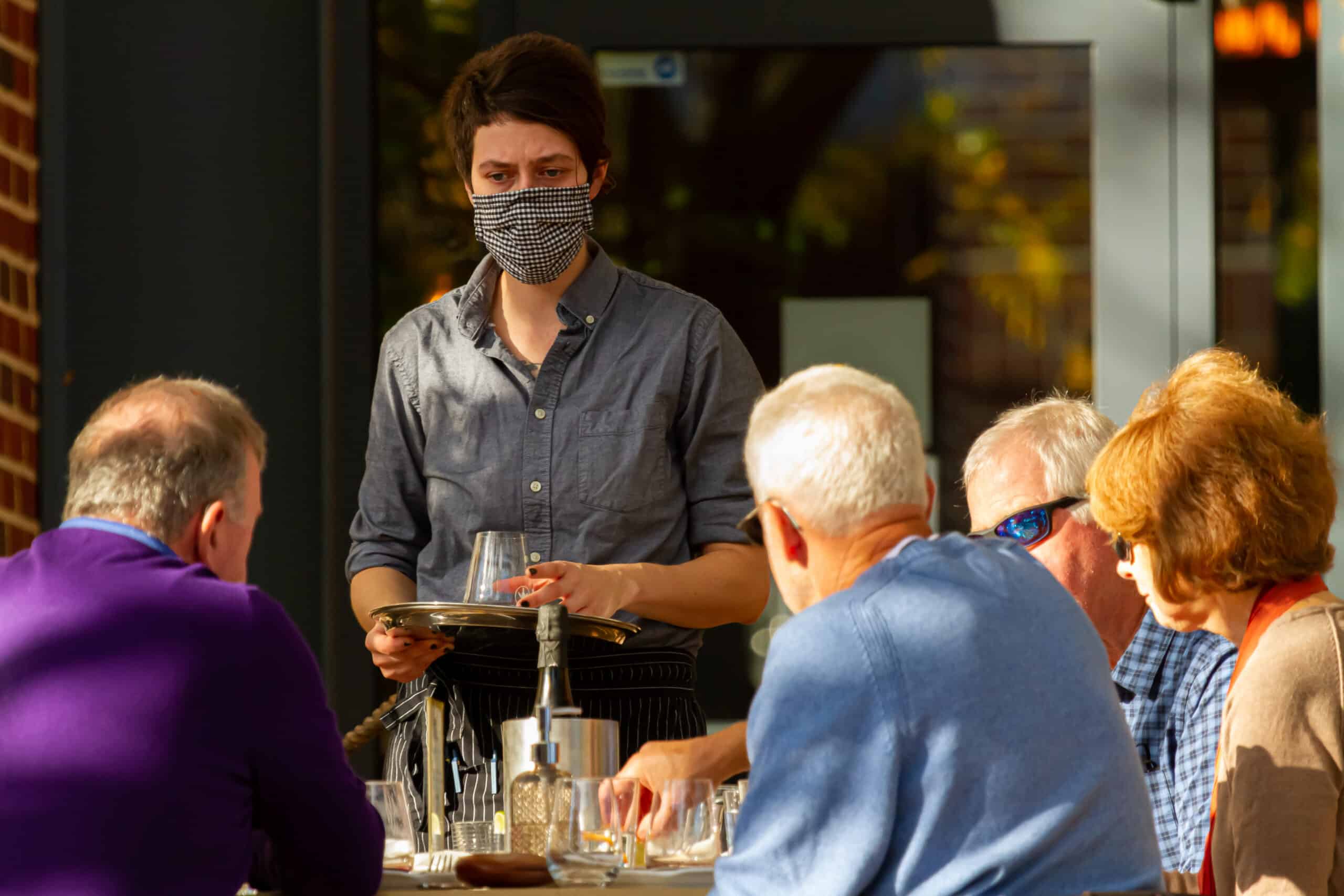 Restaurant server wearing a face mask while attending to a table of diners seated outdoors.