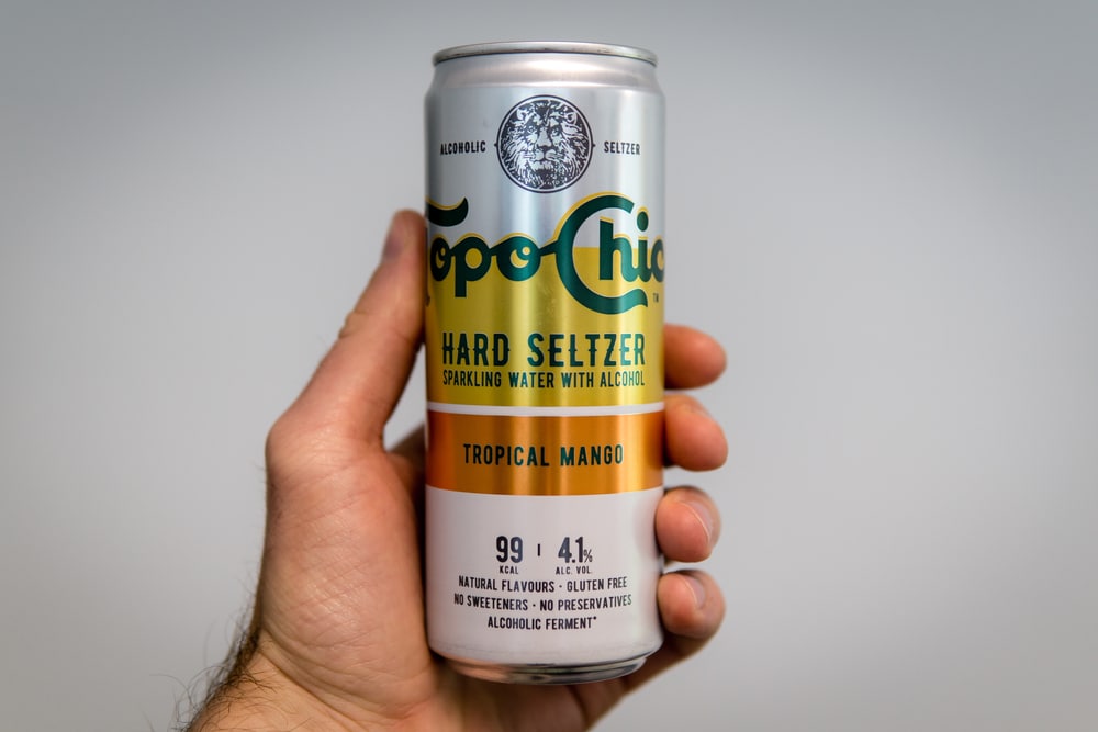 Hand holding a can of Topo Chico Tropical Mango Hard Seltzer against a plain background.