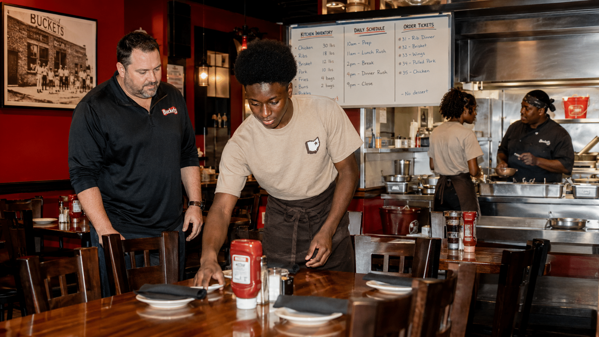 Raymel sets a dining table under a manager's guidance near the kitchen area at Buckets restaurant.