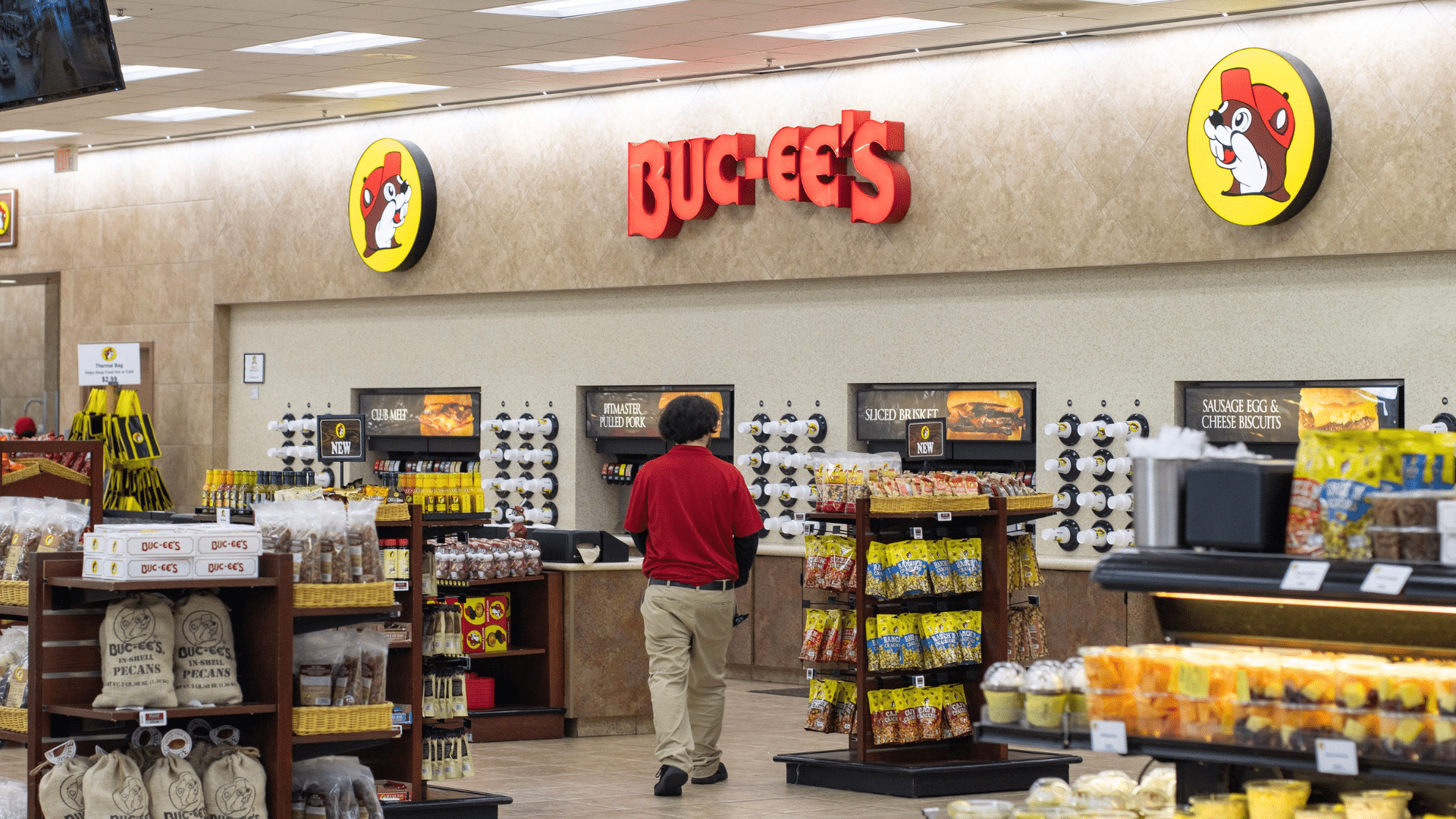 An employee in a red shirt walking past food counters and snack displays inside a brightly lit Buc-ee's store.