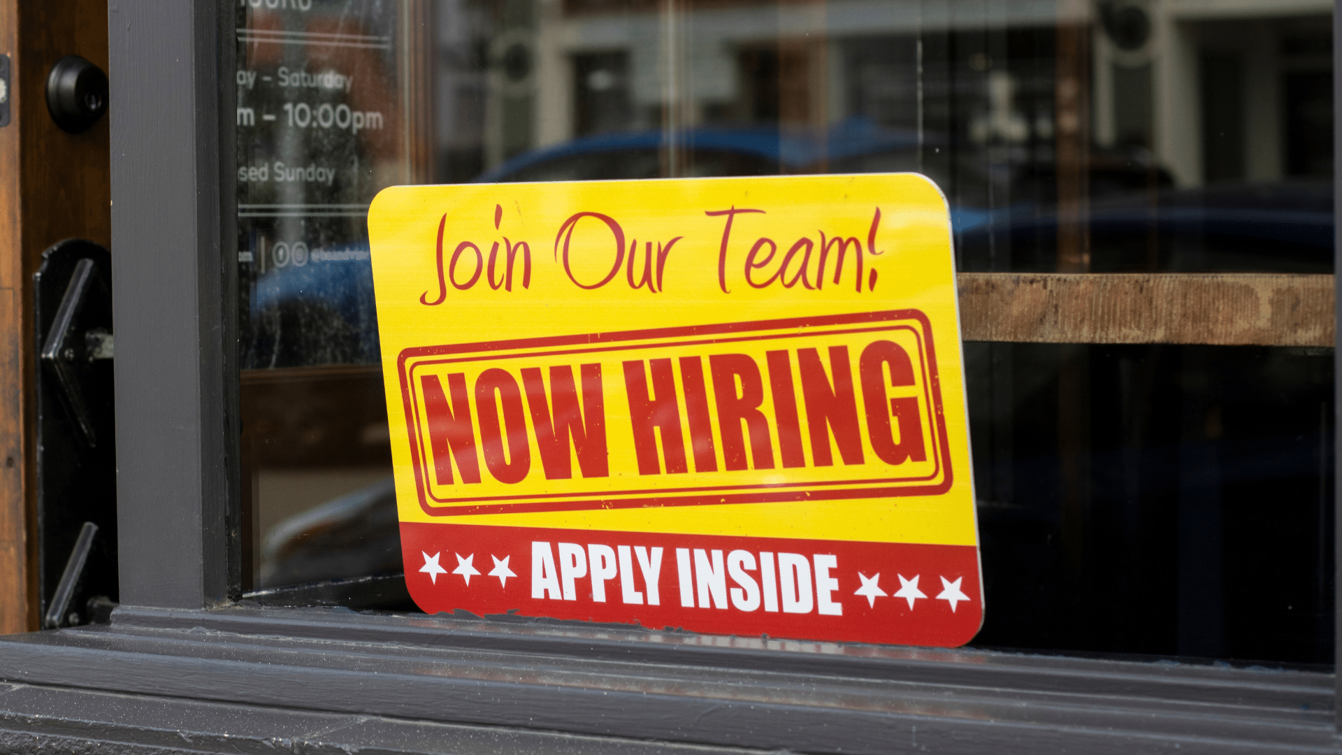 A bright yellow and red "Now Hiring" sign displayed in the glass window of a storefront.