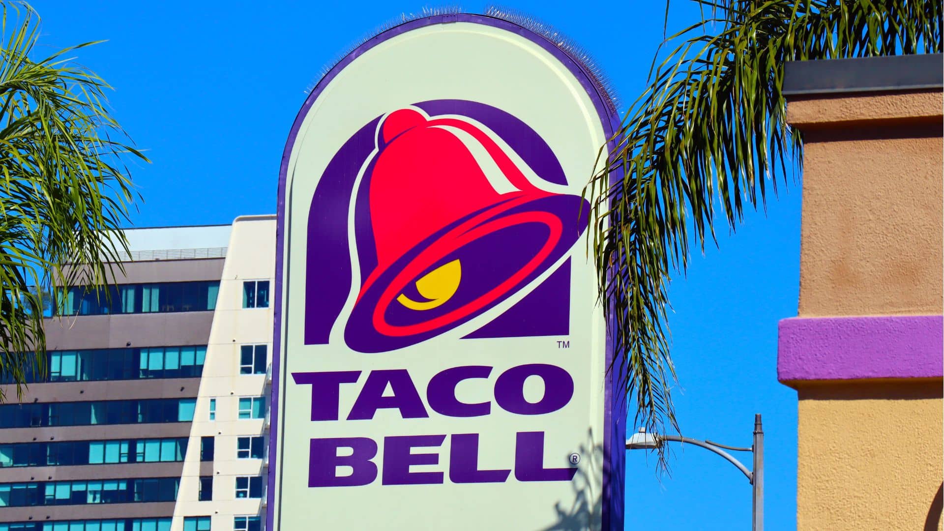 Taco Bell restaurant sign against a blue sky, with palm fronds and a high-rise building in the background.