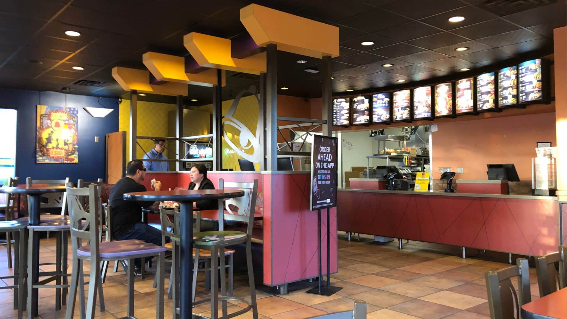Interior of a Taco Bell restaurant with customers seated at a table, an order counter, and digital menu boards in the background.
