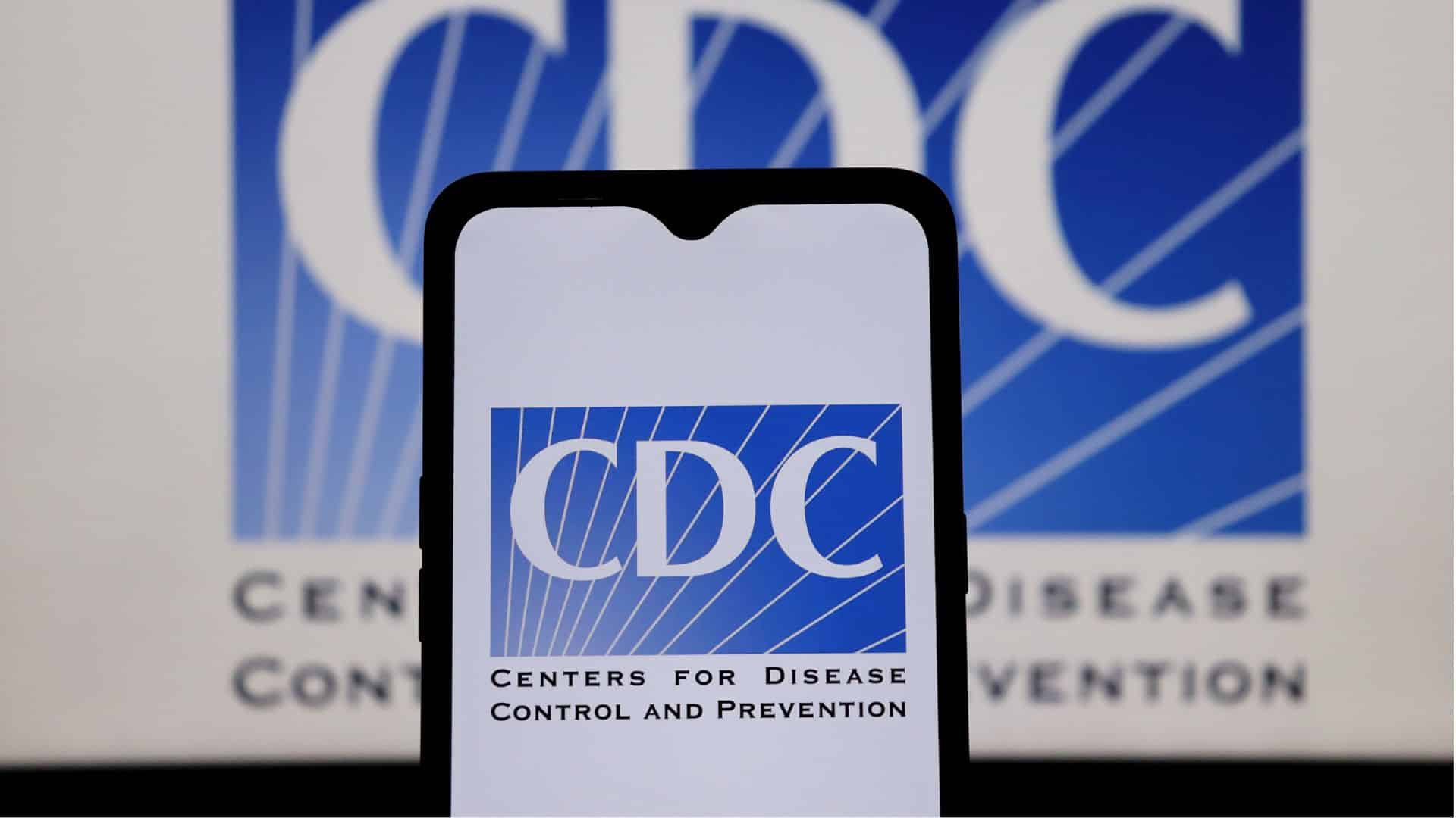 Smartphone displaying the CDC logo, held in front of a screen showing the same Centers for Disease Control and Prevention logo in the back.