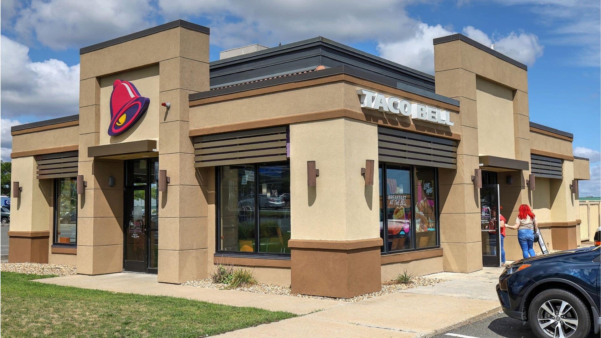 Exterior of a Taco Bell restaurant with parking lot and customers entering, under a partly cloudy sky.