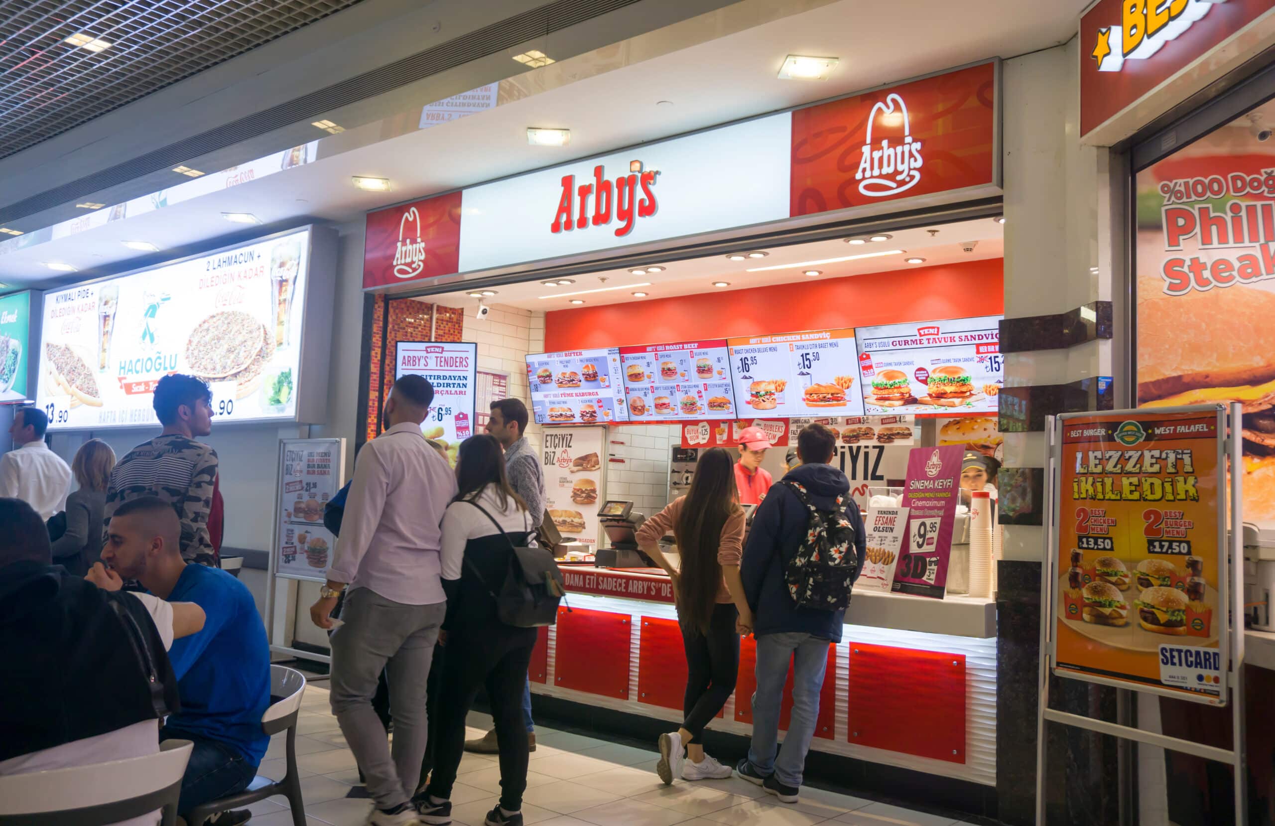 Customers ordering food at an Arby's restaurant counter inside a shopping mall food court.