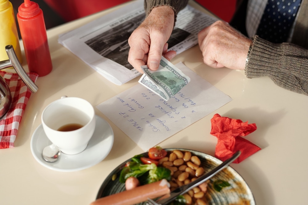 Close-up shot of man's hand holding money while preparing to pay restaurant bill next to cup of coffee and plate of food.