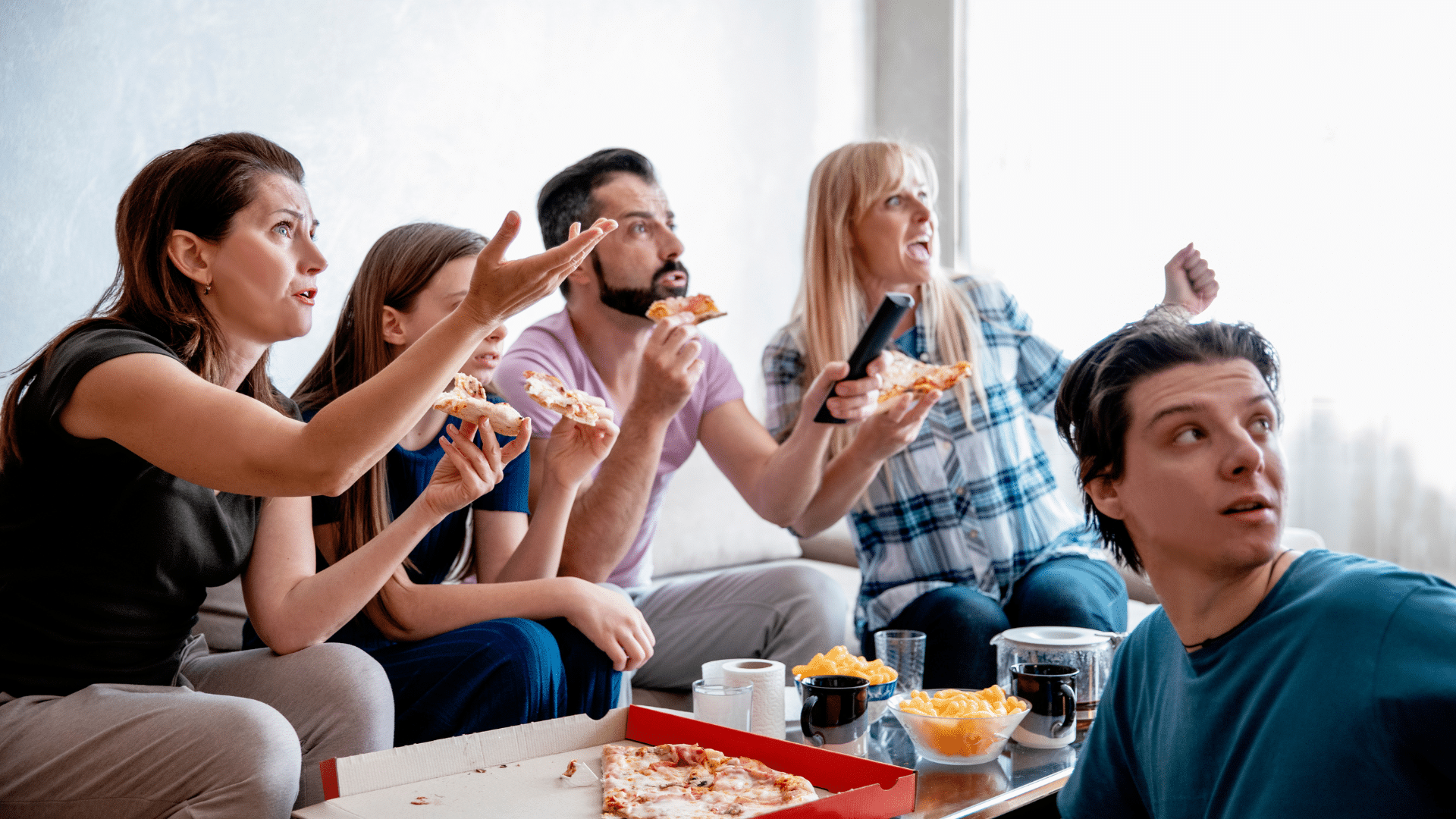 A group of friends sits on a sofa eating delivery pizza while reacting to a television program.