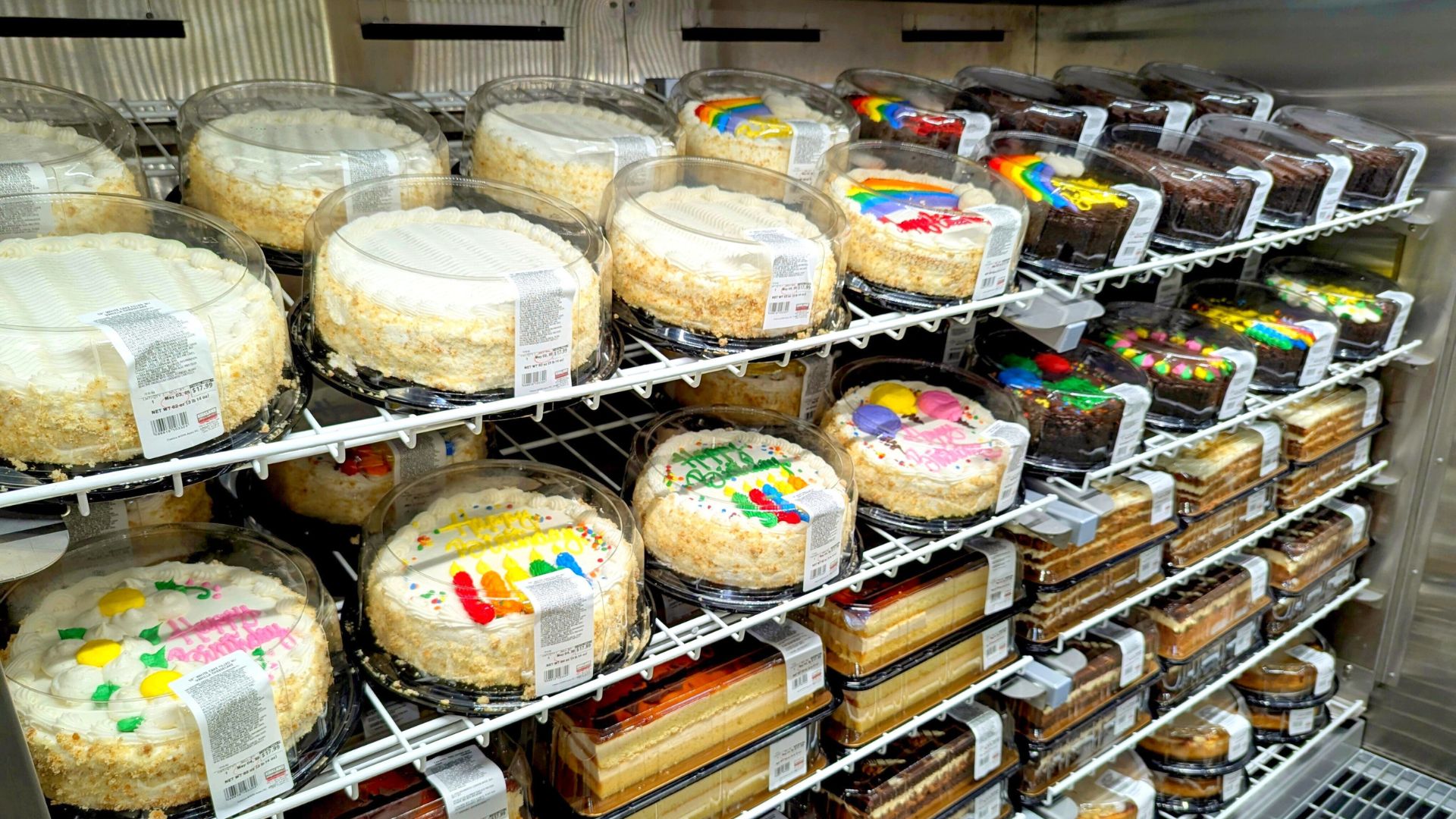 Assorted decorated cakes and desserts displayed on refrigerated store shelves.