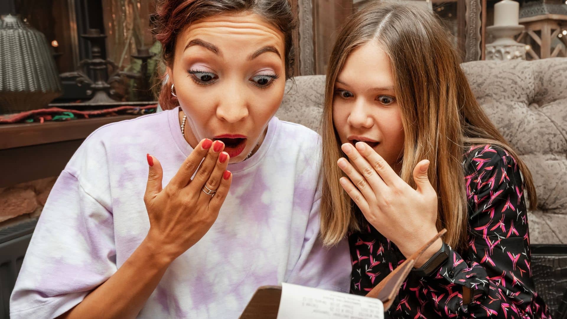 Two women reacting with surprise while looking down at a menu or bill.