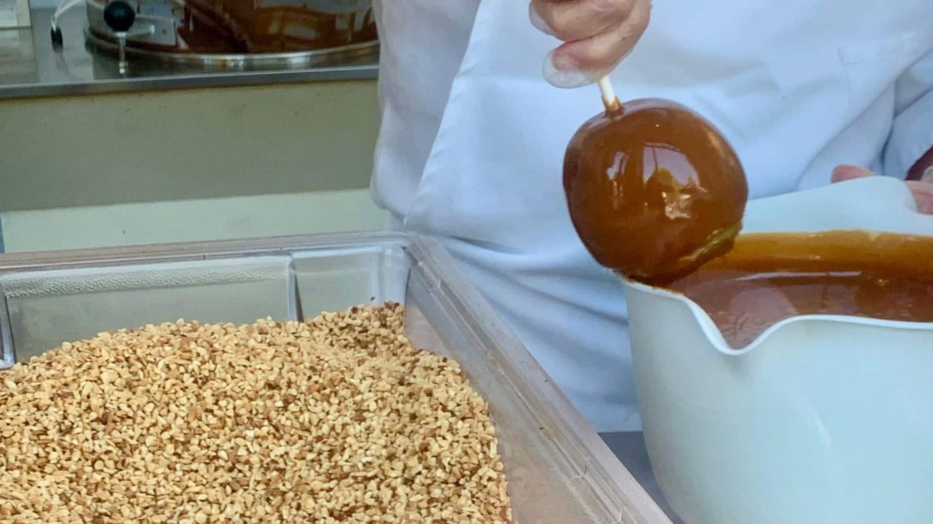 Confectioner dipping an apple into caramel beside a container of chopped nuts.