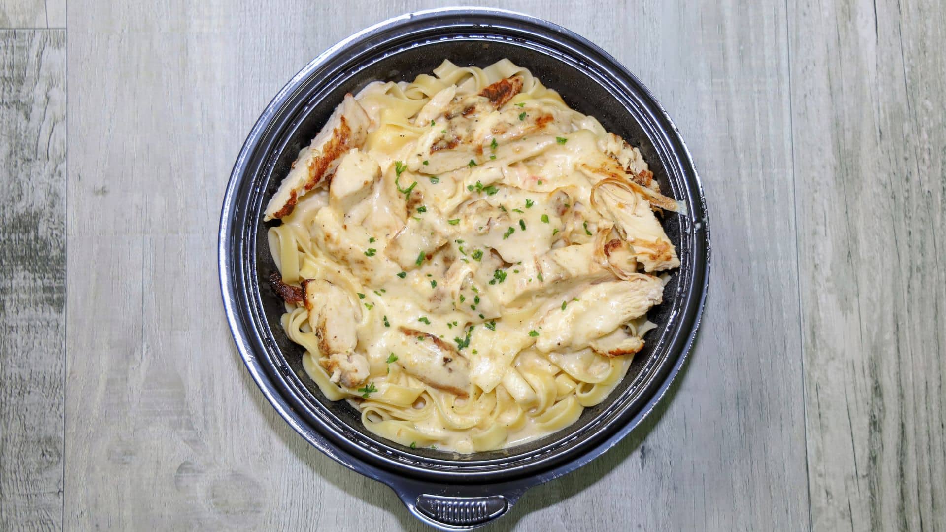 Chicken fettuccine Alfredo topped with herbs in a black takeout bowl.