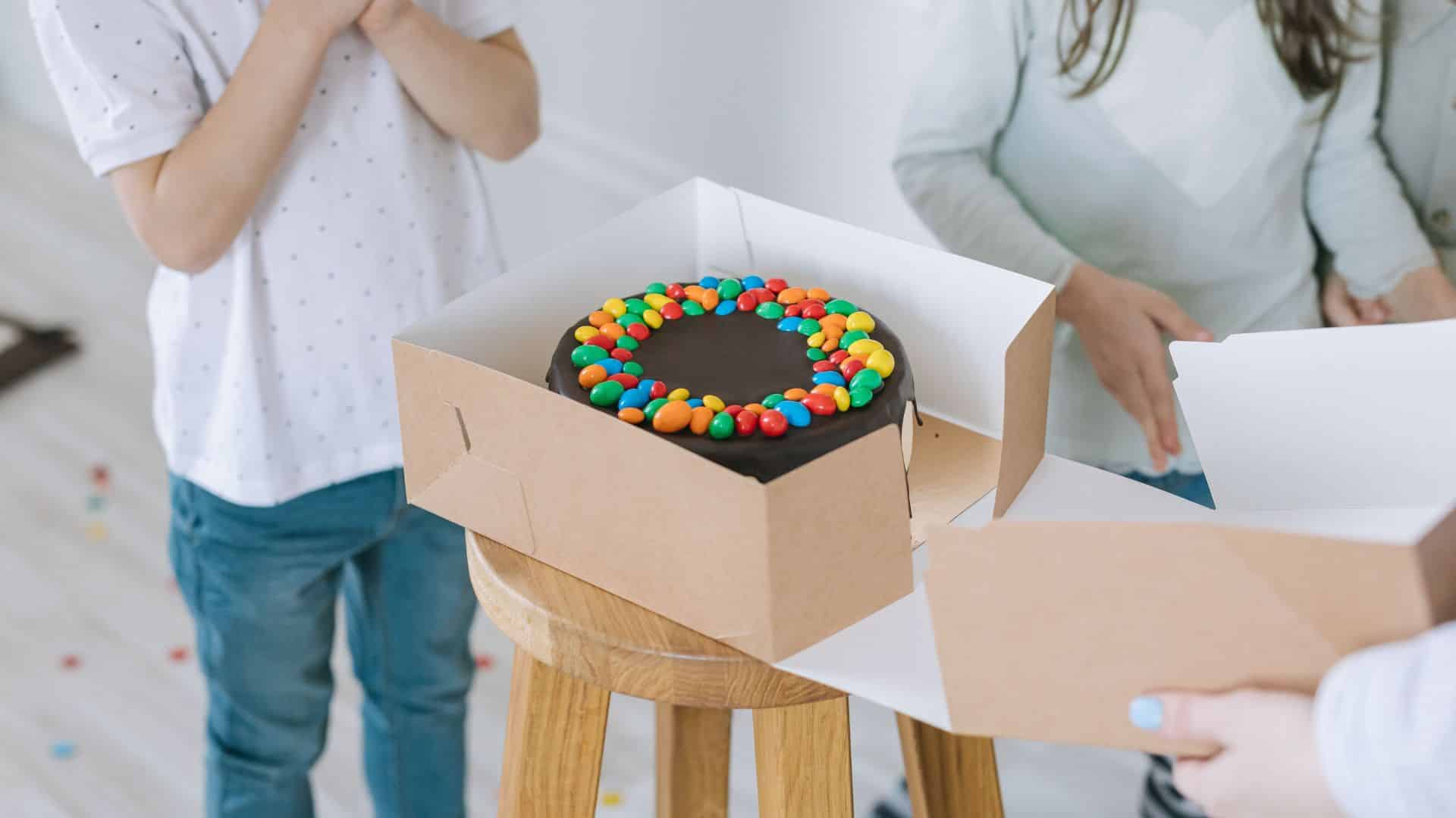 Chocolate cake topped with colorful candy pieces in an open bakery box.