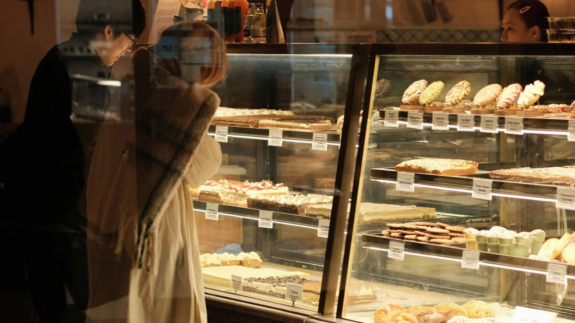 Customers browsing cakes, pastries, and other desserts in a bakery display case.