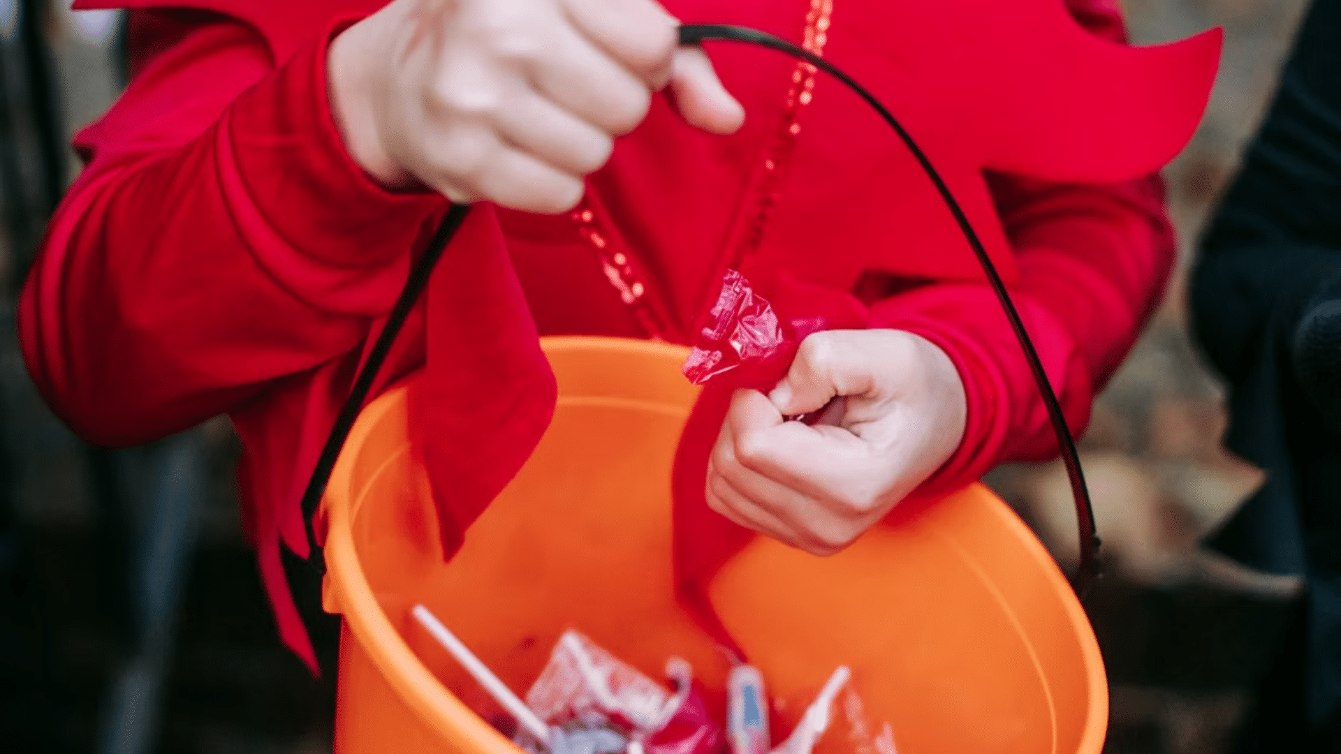A person in a red costume drops wrapped candy into an orange trick-or-treat bucket.