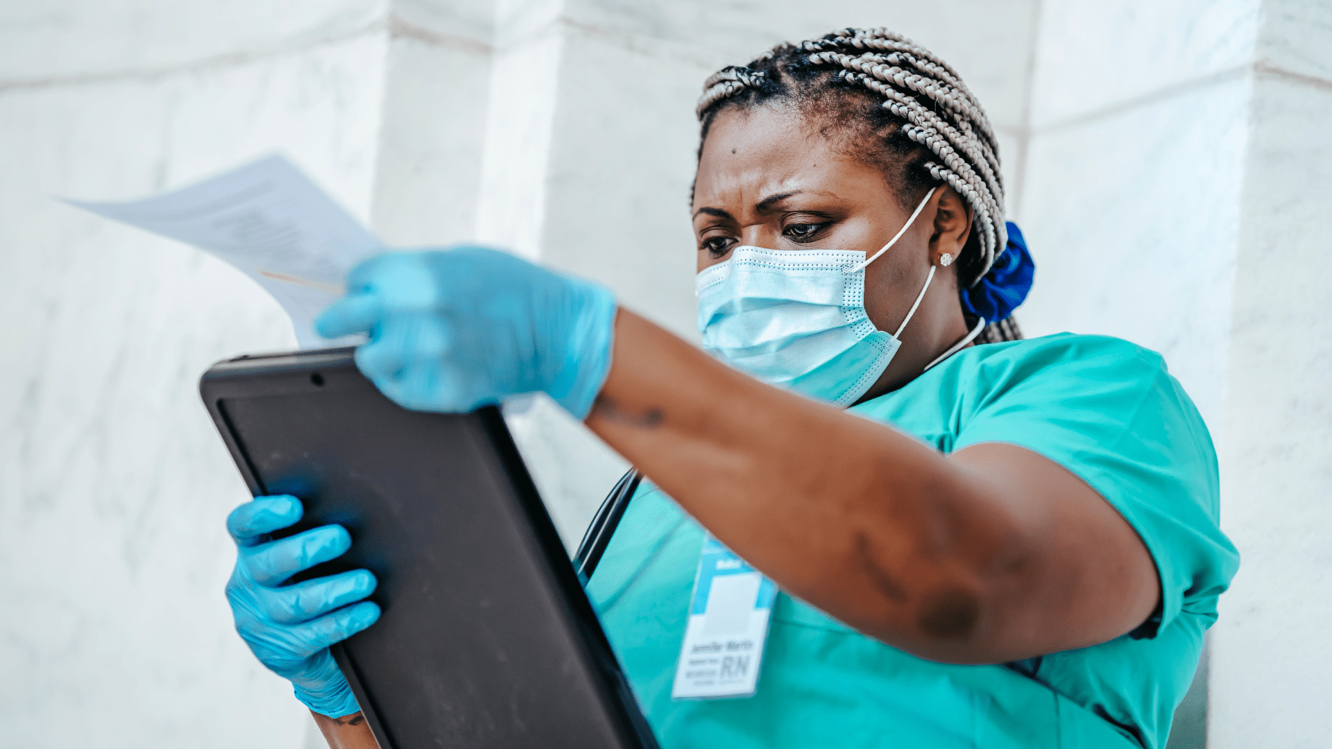 A healthcare worker wearing blue scrubs, a surgical mask, and blue latex gloves reviews documents held on a black clipboard outside against a marble wall.