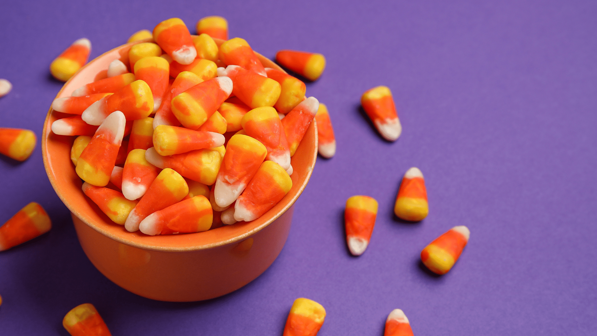 An orange bowl overflows with yellow, orange, and white candy corn pieces against a purple background.