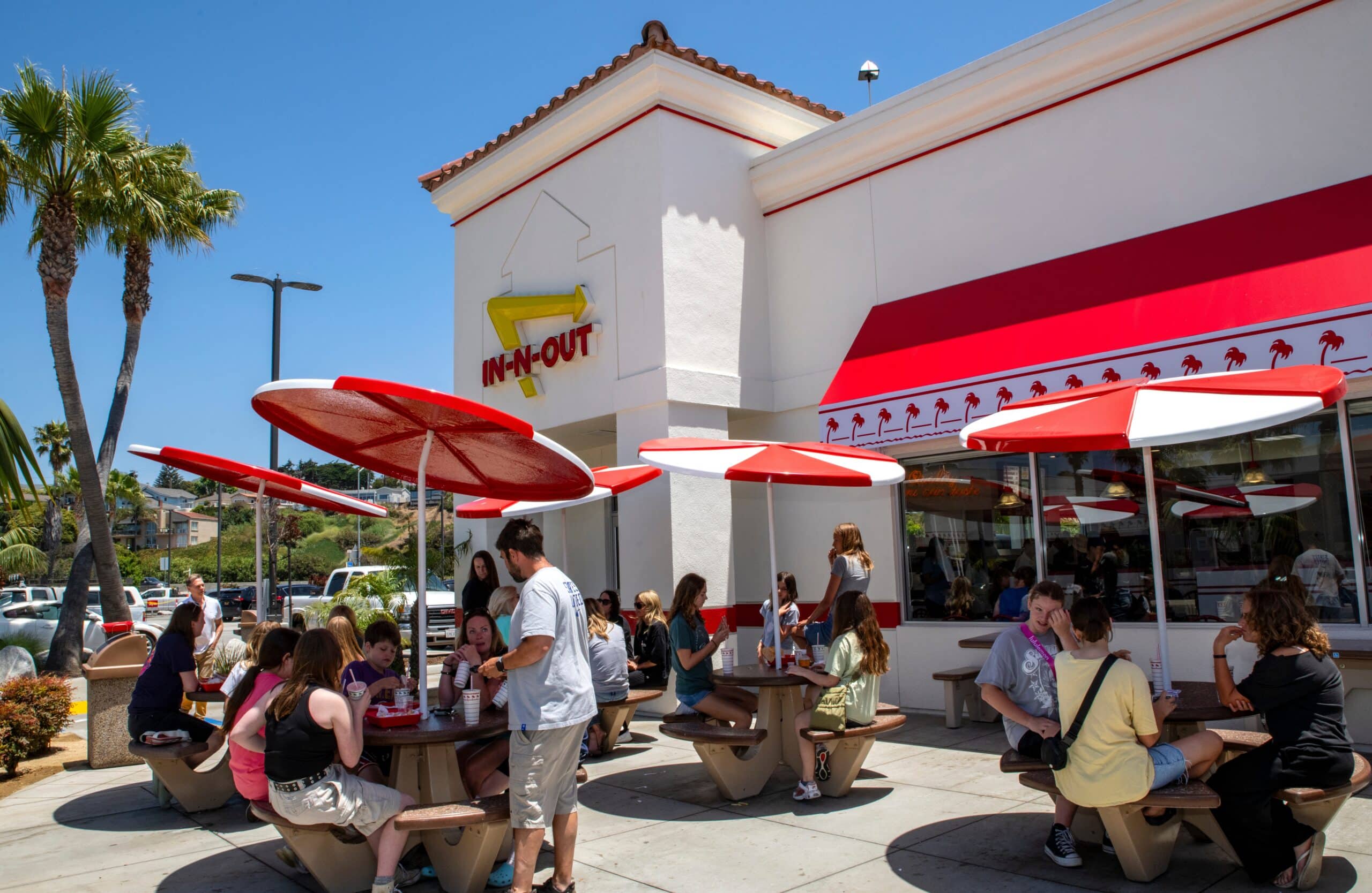 Customers eat at outdoor picnic tables beneath red-and-white umbrellas outside an In-N-Out restaurant.