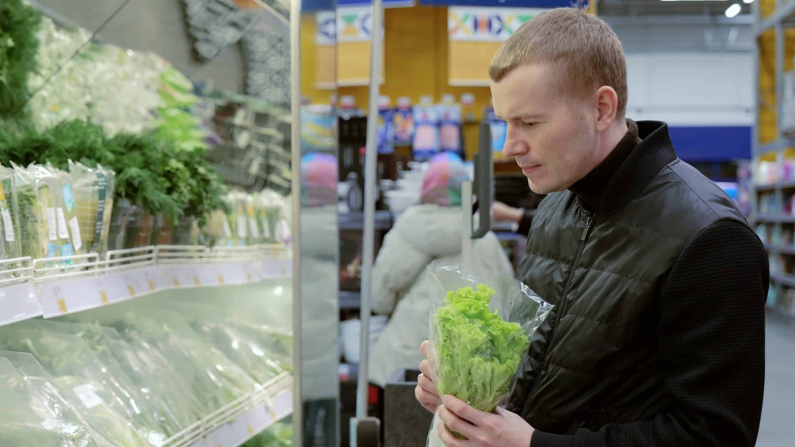 Shopper examines a bag of lettuce in a grocery store produce section while deciding what to buy.
