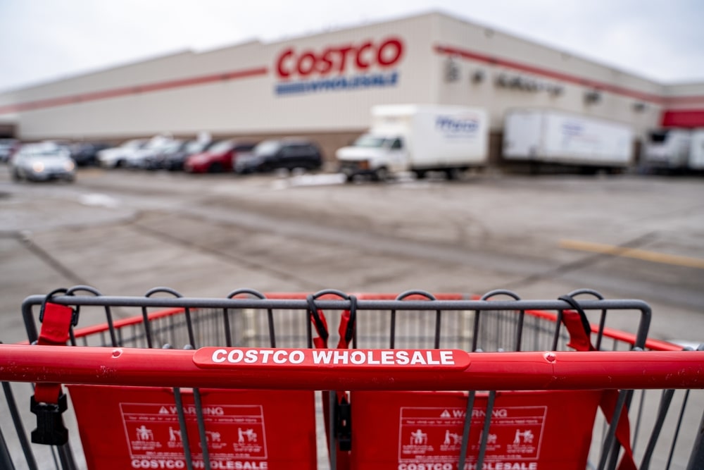 Red Costco Wholesale shopping cart in the foreground of a warehouse parking lot, with the store and delivery trucks blurred in the background.