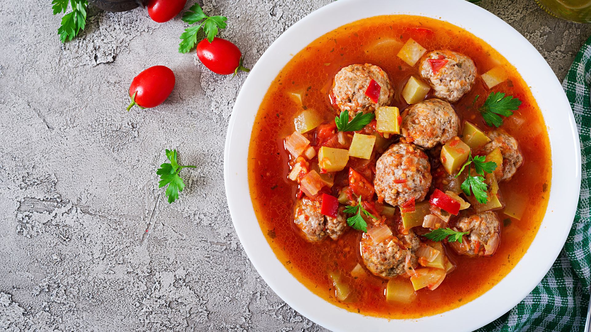 Overhead view of meatball soup with potatoes, tomatoes, peppers, and fresh parsley in a white bowl.