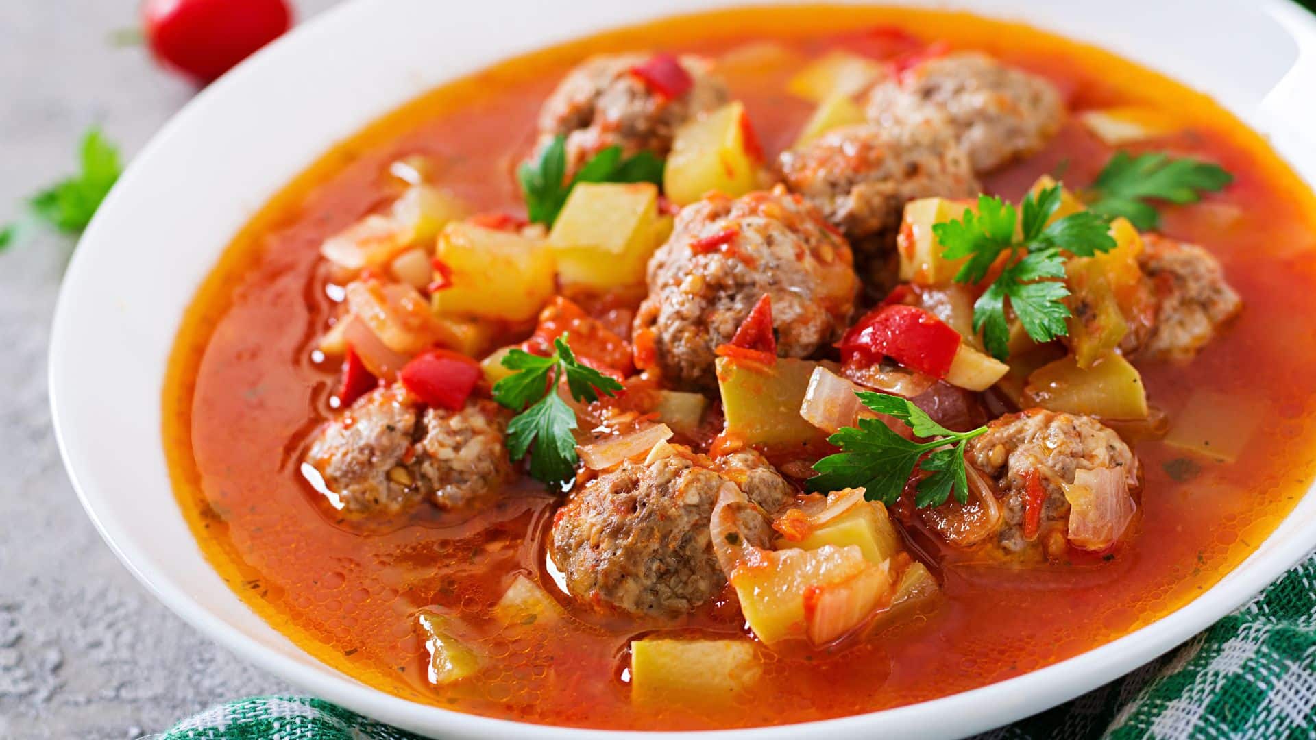 Close-up of meatball soup in a tomato-based broth with diced vegetables and fresh parsley garnish.