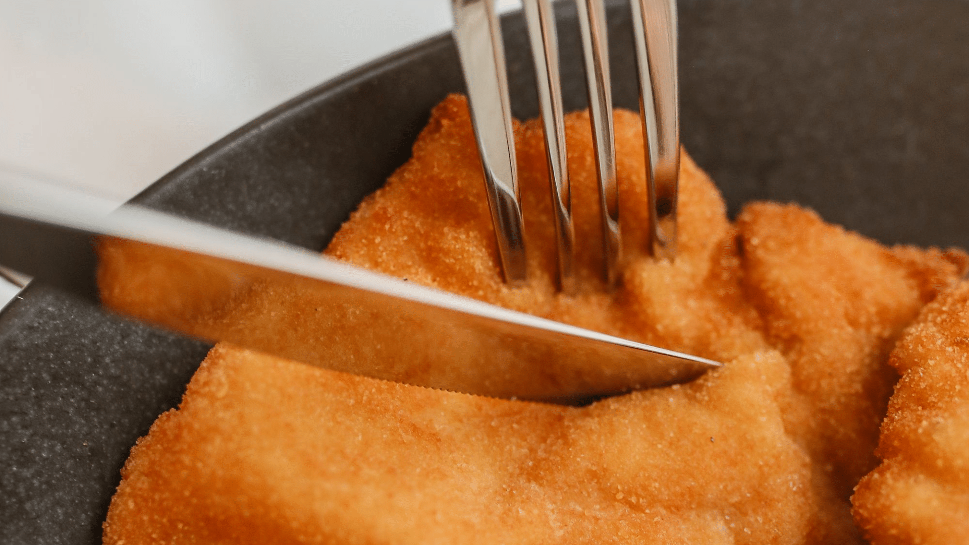 A close-up shot shows a silver fork pressing down into a crispy, golden-brown schnitzel as a knife begins to cut through it.