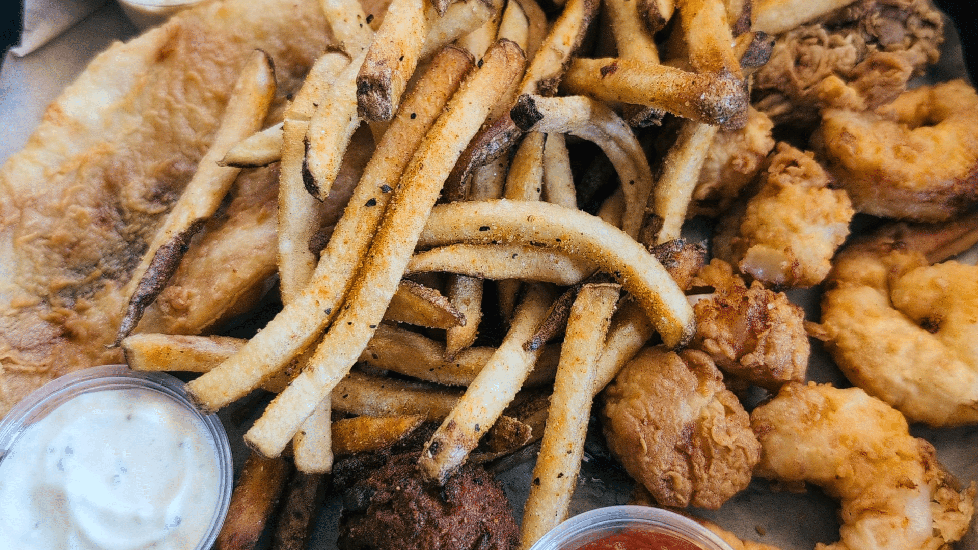 A round platter filled with fried seafood, French fries, hush puppies, coleslaw, and small plastic cups of dipping sauces.