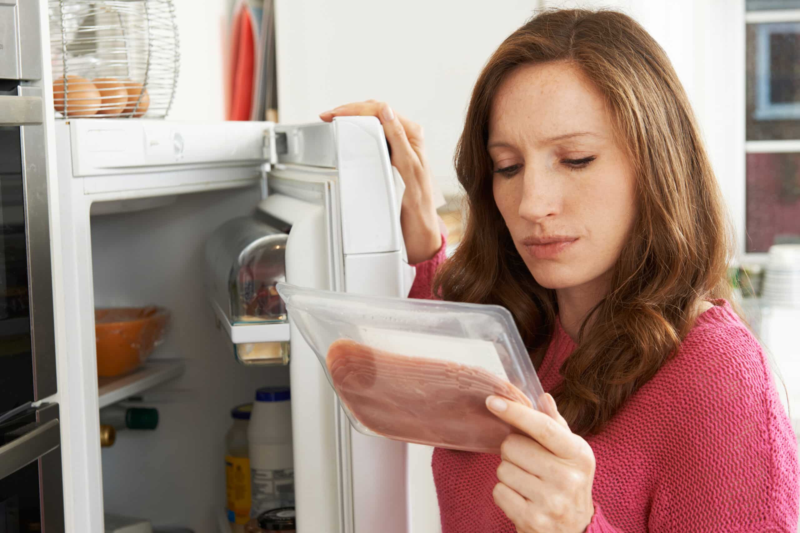 Woman examining a package of deli meat while standing in front of an open refrigerator at home.